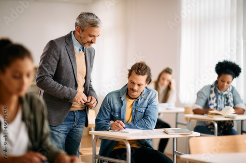 Smiling mature professor assists his student with lecture during class in classroom.