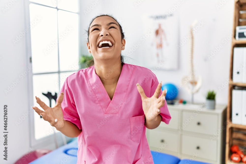 Obraz premium Young hispanic woman working at pain recovery clinic crazy and mad shouting and yelling with aggressive expression and arms raised. frustration concept.