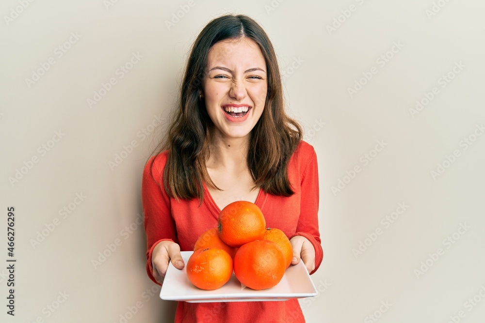 Young brunette woman holding plate with fresh oranges smiling and laughing hard out loud because funny crazy joke. Young brunette woman holding plate with fresh oranges smiling and laughing hard out loud because funny crazy joke.