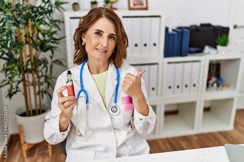 Middle age doctor woman holding electronic cigarette at medical clinic smiling happy and positive, thumb up doing excellent and approval sign