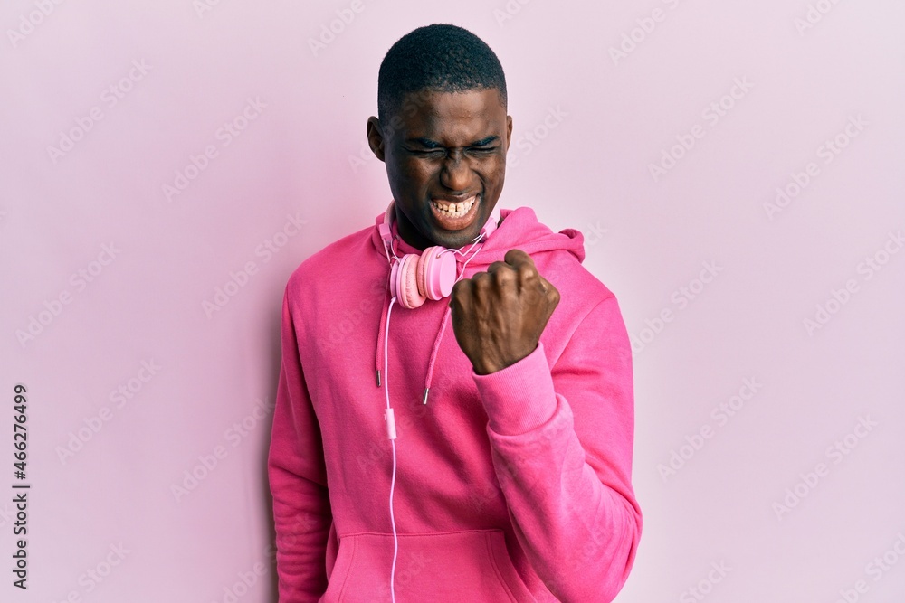 Young african american man wearing gym clothes and using headphones angry and mad raising fist frustrated and furious while shouting with anger. rage and aggressive concept. Young african american man wearing gym clothes and using headphones angry and mad raising fist frustrated and furious while shouting with anger. rage and aggressive concept.