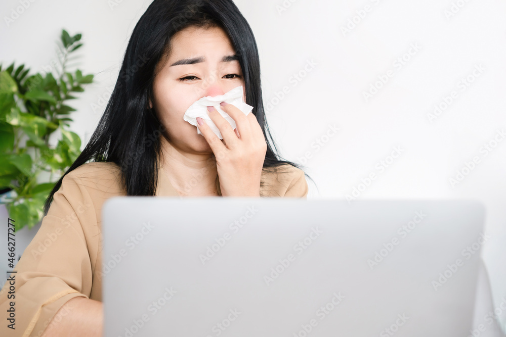 unwell Asian woman sneezing and coughing because of flu and fever while working on computer at desk hand holding tissue papers unwell Asian woman sneezing and coughing because of flu and fever while working on computer at desk hand holding tissue papers