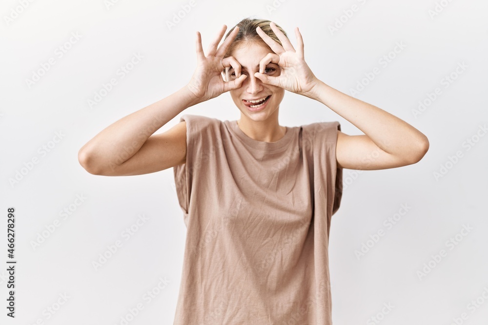 Young blonde woman standing over isolated background doing ok gesture like binoculars sticking tongue out, eyes looking through fingers. crazy expression. Young blonde woman standing over isolated background doing ok gesture like binoculars sticking tongue out, eyes looking through fingers. crazy expression.