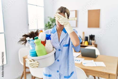 Young blonde woman wearing cleaner uniform holding cleaning products covering eyes with hand  looking serious and sad. sightless  hiding and rejection concept