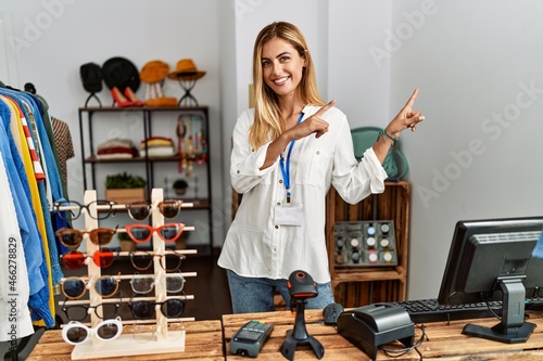 Blonde beautiful young woman working as manager at retail boutique smiling and looking at the camera pointing with two hands and fingers to the side.