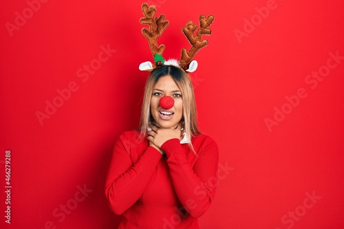 Beautiful hispanic woman wearing deer christmas hat and red nose shouting and suffocate because painful strangle. health problem. asphyxiate and suicide concept.