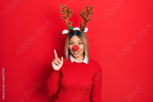 Beautiful hispanic woman wearing deer christmas hat and red nose showing and pointing up with finger number one while smiling confident and happy.
