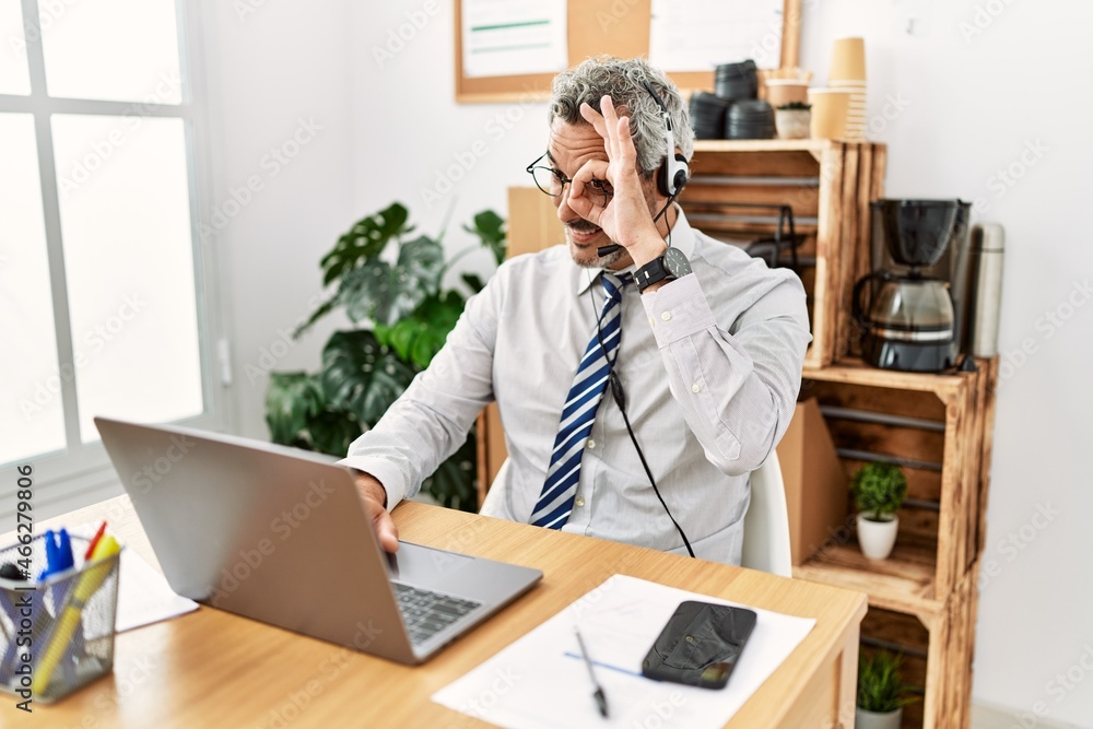 Middle age hispanic business man working at the office wearing operator headset doing ok gesture with hand smiling, eye looking through fingers with happy face. Middle age hispanic business man working at the office wearing operator headset doing ok gesture with hand smiling, eye looking through fingers with happy face.