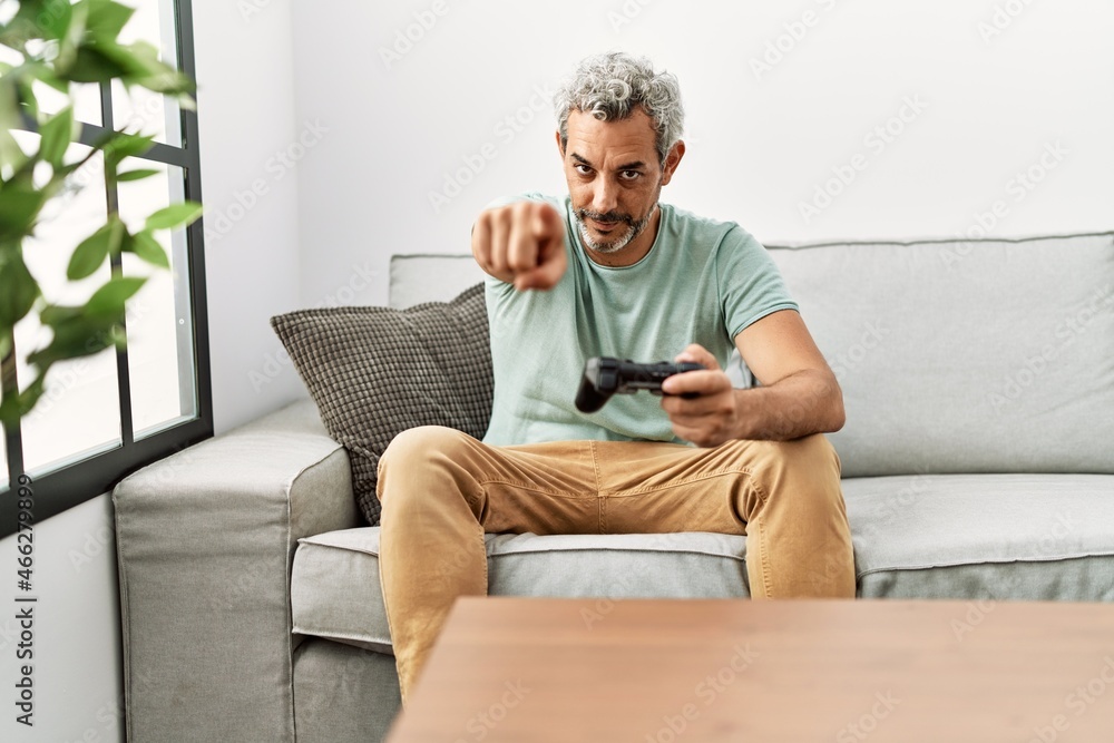 Middle age hispanic man playing video game sitting on the sofa pointing with finger to the camera and to you, confident gesture looking serious