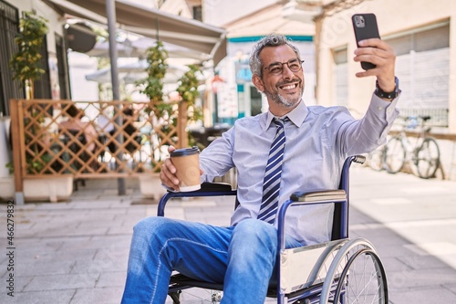 Middle age hispanic man sitting on wheelchair makes selfie by the smartphone at street