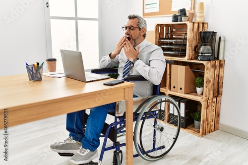 Middle age hispanic man working at the office sitting on wheelchair bored yawning tired covering mouth with hand. restless and sleepiness.