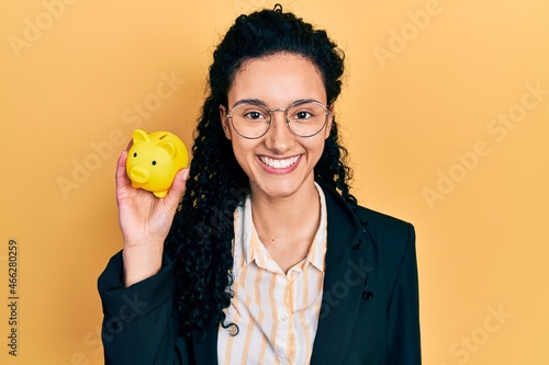Young hispanic woman with curly hair holding piggy bank looking positive and happy standing and smiling with a confident smile showing teeth
