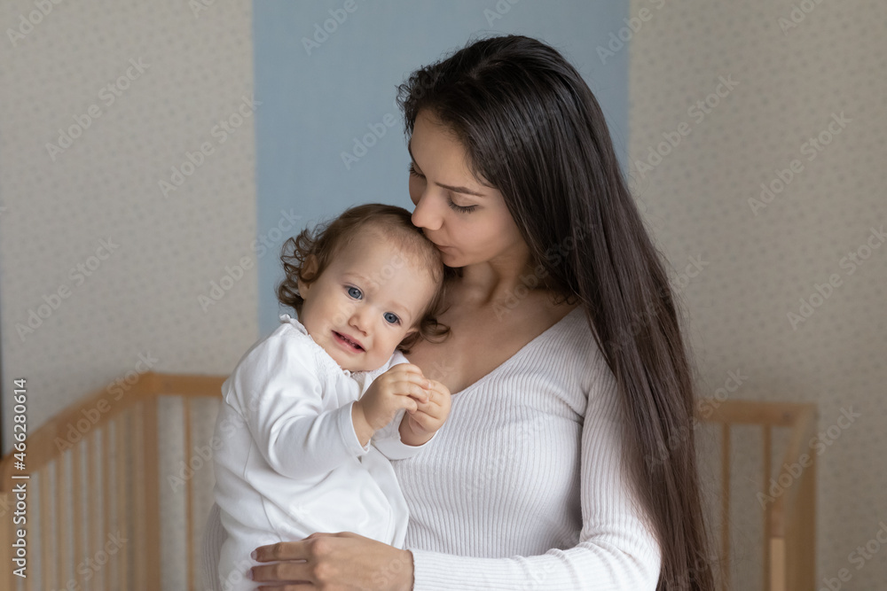 Loving young mother kissing little daughter on forehead, caring mom holding hugging adorable toddler child kid, family enjoying tender moment at home in nursery, motherhood and childcare concept Loving young mother kissing little daughter on forehead, caring mom holding hugging adorable toddler child kid, family enjoying tender moment at home in nursery, motherhood and childcare concept