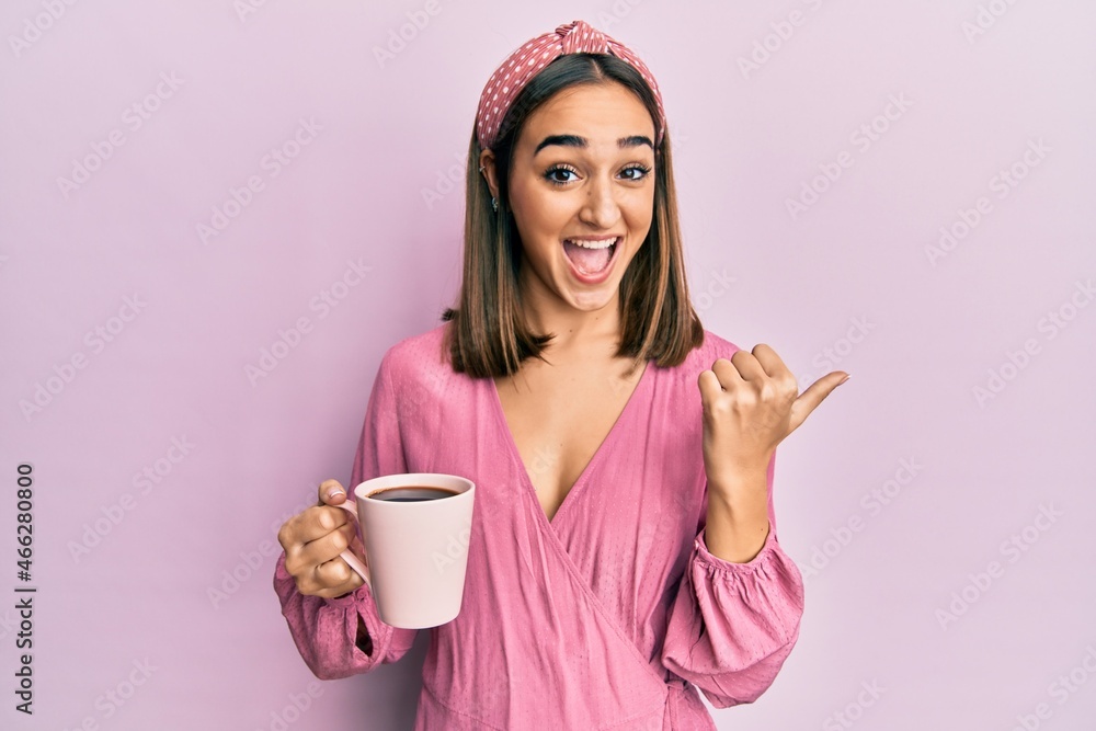 Young brunette girl drinking a cup of coffee pointing thumb up to the side smiling happy with open mouth Young brunette girl drinking a cup of coffee pointing thumb up to the side smiling happy with open mouth