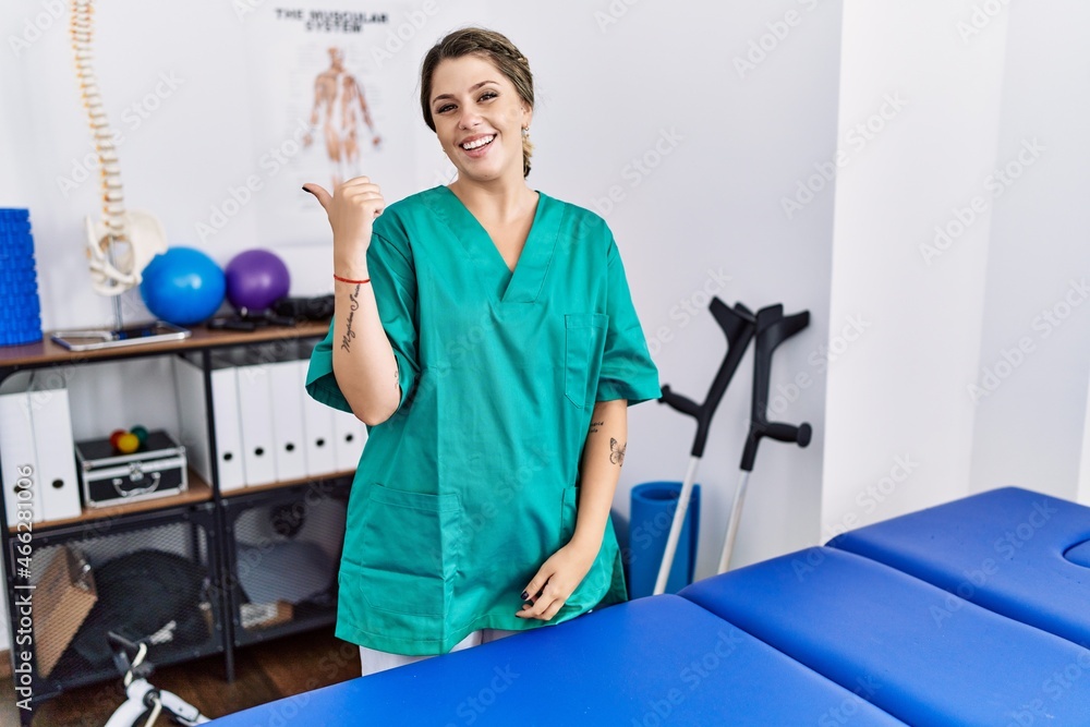 Young hispanic woman wearing physiotherapist uniform standing at clinic smiling with happy face looking and pointing to the side with thumb up. Young hispanic woman wearing physiotherapist uniform standing at clinic smiling with happy face looking and pointing to the side with thumb up.