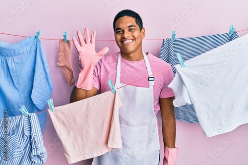 Young handsome hispanic man wearing cleaner apron holding clothes on clothesline showing and pointing up with fingers number five while smiling confident and happy.
