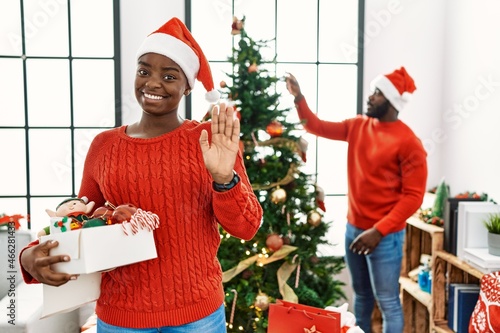 Young african american couple standing by christmas tree waiving saying hello happy and smiling, friendly welcome gesture