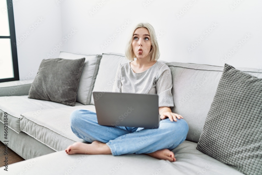 Young caucasian woman using laptop at home sitting on the sofa making fish face with lips, crazy and comical gesture. funny expression. Young caucasian woman using laptop at home sitting on the sofa making fish face with lips, crazy and comical gesture. funny expression.
