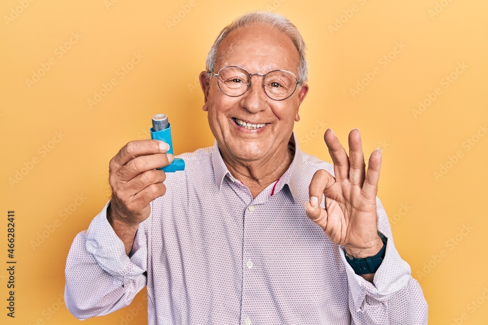 Senior man with grey hair holding medical asthma inhaler doing ok sign with fingers, smiling friendly gesturing excellent symbol - obrazy, fototapety, plakaty 