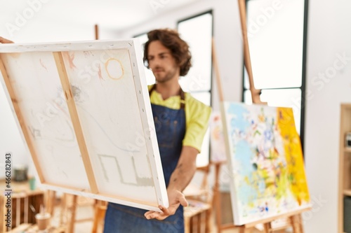 Young hispanic artist man concentrate holding and looking to canvas at art studio.