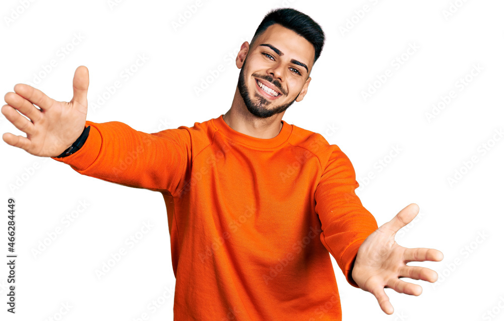 Young hispanic man with beard wearing casual orange sweater looking at the camera smiling with open arms for hug. cheerful expression embracing happiness. Young hispanic man with beard wearing casual orange sweater looking at the camera smiling with open arms for hug. cheerful expression embracing happiness.
