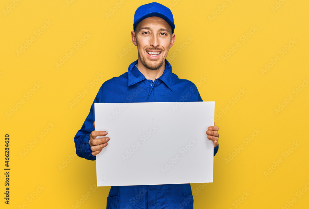 Bald man with beard wearing builder jumpsuit uniform holding empty banner looking positive and happy standing and smiling with a confident smile showing teeth Bald man with beard wearing builder jumpsuit uniform holding empty banner looking positive and happy standing and smiling with a confident smile showing teeth