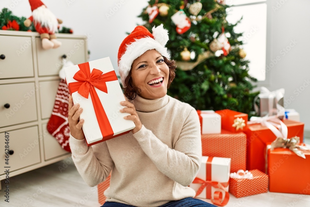 Middle age hispanic woman holding gift sitting by christmas tree at home. Middle age hispanic woman holding gift sitting by christmas tree at home.