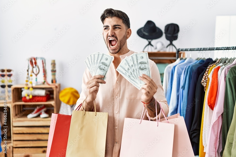 Young hispanic man holding shopping bags and dollars at retail shop angry and mad screaming frustrated and furious, shouting with anger. rage and aggressive concept. Young hispanic man holding shopping bags and dollars at retail shop angry and mad screaming frustrated and furious, shouting with anger. rage and aggressive concept.