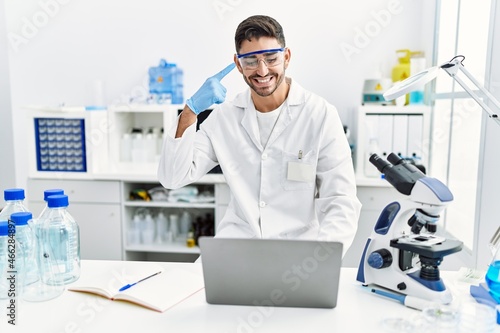 Young hispanic man working at scientist laboratory with laptop smiling happy pointing with hand and finger