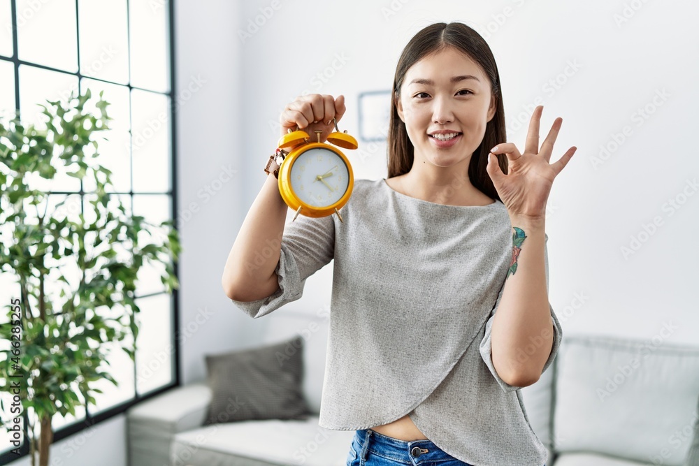Young asian woman holding alarm clock doing ok sign with fingers, smiling friendly gesturing excellent symbol Young asian woman holding alarm clock doing ok sign with fingers, smiling friendly gesturing excellent symbol