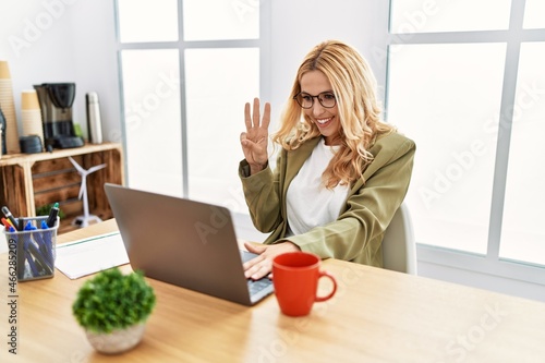Beautiful blonde woman working at the office with laptop showing and pointing up with fingers number three while smiling confident and happy.