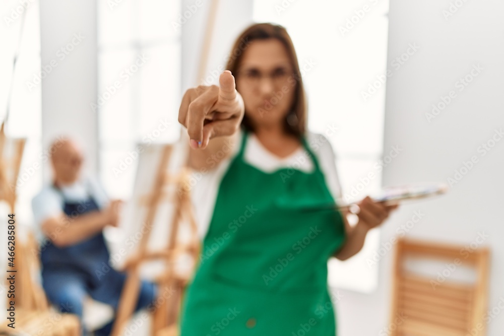 Middle age hispanic woman at art studio pointing with finger to the camera and to you, confident gesture looking serious Middle age hispanic woman at art studio pointing with finger to the camera and to you, confident gesture looking serious