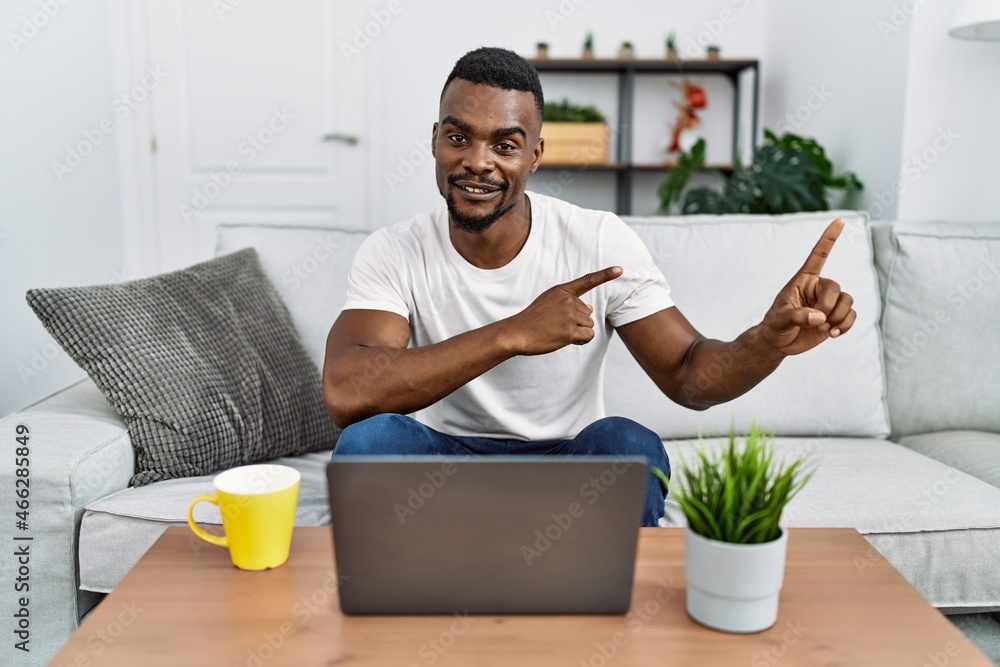 Young african man using laptop at home smiling and looking at the camera pointing with two hands and fingers to the side. Young african man using laptop at home smiling and looking at the camera pointing with two hands and fingers to the side.
