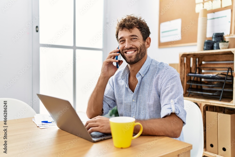 Young handsome man working at the office speaking on the phone looking positive and happy standing and smiling with a confident smile showing teeth Young handsome man working at the office speaking on the phone looking positive and happy standing and smiling with a confident smile showing teeth