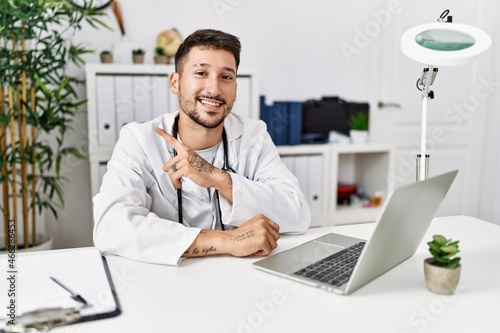 Young doctor working at the clinic using computer laptop with a big smile on face, pointing with hand and finger to the side looking at the camera.