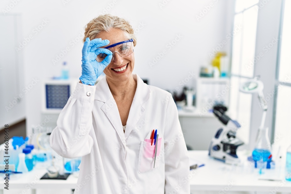 Middle age blonde woman working at scientist laboratory doing ok gesture with hand smiling, eye looking through fingers with happy face.