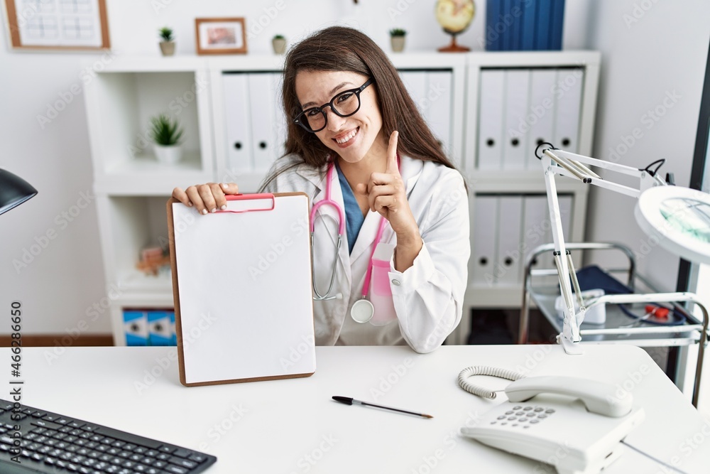 Young doctor woman holding clipboard at the clinic smiling with an idea or question pointing finger with happy face, number one Young doctor woman holding clipboard at the clinic smiling with an idea or question pointing finger with happy face, number one