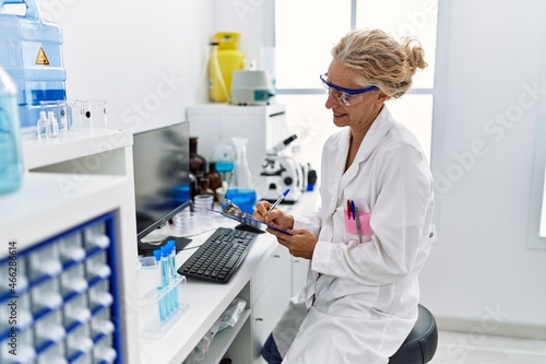 Middle age blonde woman wearing scientist uniform writing on clipboard working at laboratory