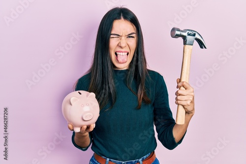 Young brunette woman holding piggy bank and hammer sticking tongue out happy with funny expression.