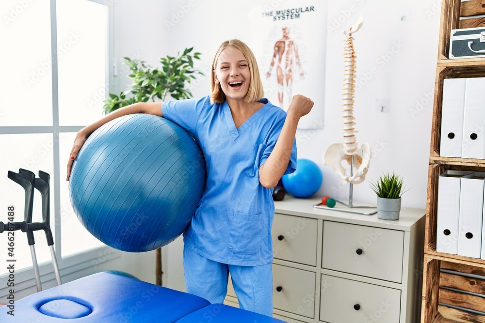 Young caucasian physiotherapist woman holding pilates ball screaming proud, celebrating victory and success very excited with raised arms