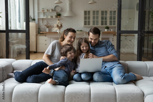 Full length happy young parents and adorable little children siblings watching funny movie or cartoons online on computer, relaxing on comfortable couch in modern living room, spending weekend at home
