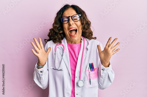Middle age hispanic woman wearing doctor uniform and glasses celebrating mad and crazy for success with arms raised and closed eyes screaming excited. winner concept