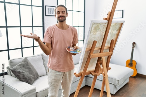 Young hispanic man with beard painting on canvas at home smiling cheerful presenting and pointing with palm of hand looking at the camera.