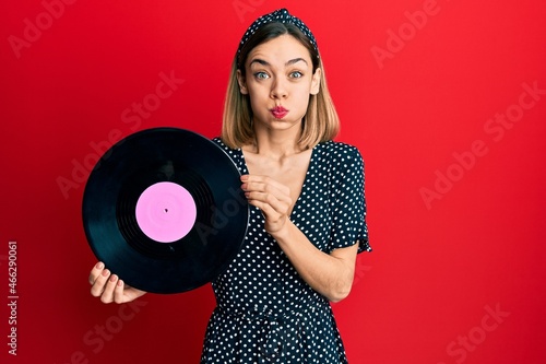 Young caucasian blonde woman holding vinyl disc puffing cheeks with funny face. mouth inflated with air, catching air.