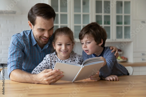 Happy young father reading paper book to little adorable kids son daughter, telling interesting fairy tale story together in modern kitchen, enjoying leisure weekend pastime hobby activity indoors.