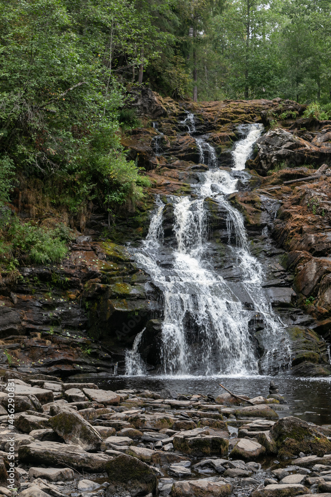 Fototapeta premium A high waterfall in the middle of a green northern forest. The rocks around the waterfall are rugged and create many steps. Water foams and flows as a river through the forest along the stones.