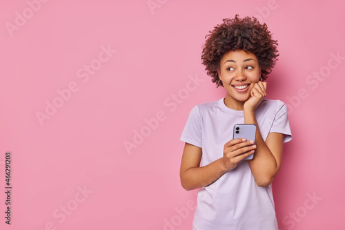 Horizontal shot of positive curly haired woman uses mobile phone downloads new application for chatting online keeps hand on face wears casual t shirt isolated over pink backgroud blank space