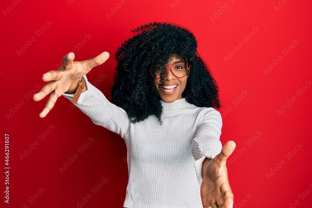 African american woman with afro hair wearing casual sweater and glasses looking at the camera smiling with open arms for hug. cheerful expression embracing happiness. African american woman with afro hair wearing casual sweater and glasses looking at the camera smiling with open arms for hug. cheerful expression embracing happiness.