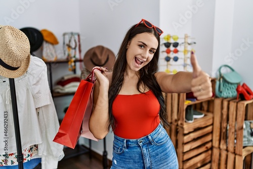 Young brunette woman holding shopping bags at retail shop approving doing positive gesture with hand, thumbs up smiling and happy for success. winner gesture.