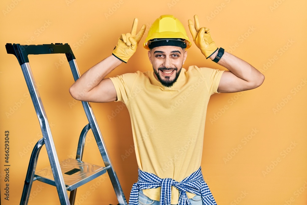 Handsome man with beard by construction stairs wearing hardhat posing funny and crazy with fingers on head as bunny ears, smiling cheerful Handsome man with beard by construction stairs wearing hardhat posing funny and crazy with fingers on head as bunny ears, smiling cheerful